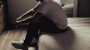 A man sits thoughtfully on the edge of a bed in a dimly lit bedroom, evoking introspection.