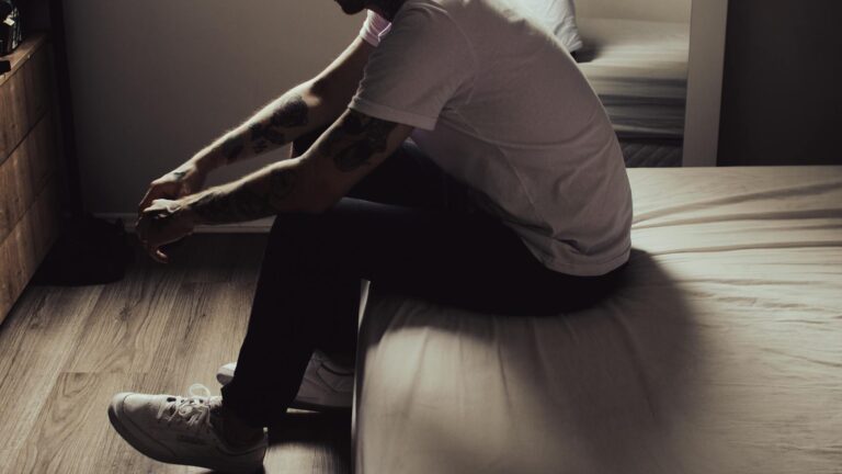 A man sits thoughtfully on the edge of a bed in a dimly lit bedroom, evoking introspection.