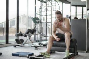Tired young muscular male athlete in casual sportswear and sneakers sitting on bench and resting after intense weightlifting workout in modern fitness club