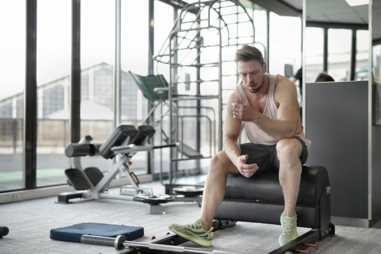 Tired young muscular male athlete in casual sportswear and sneakers sitting on bench and resting after intense weightlifting workout in modern fitness club