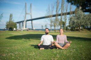 A couple practicing yoga in the park with a famous bridge in the background, capturing a moment of relaxation and mindfulness.