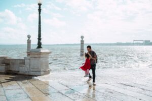 A father joyfully swings his daughter by the sea on a bright day, capturing a playful moment.
