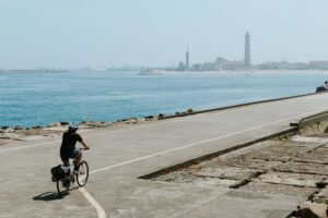 Person cycling along a coastal path with lighthouse and cityscape in view.