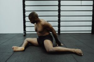 A shirtless man stretches on a black mat indoors, focusing on flexibility and fitness.