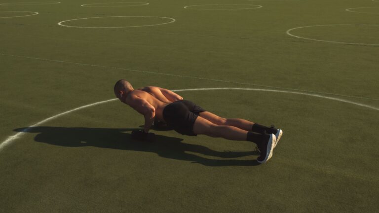 A shirtless man performing push-ups on a green field under bright daylight.