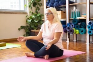 Adult woman sits cross-legged on a mat meditating in a serene yoga studio environment.