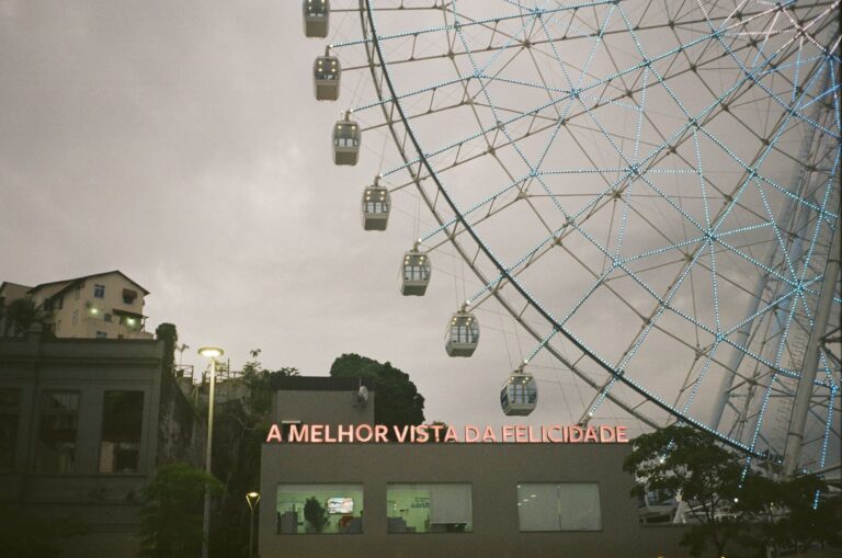 From below of Ferris wheel with cabins and modern buildings against cloudless sky