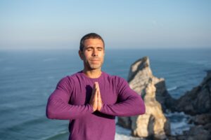 Man meditating with hands together by a rocky coastline in Portugal, ocean in background.
