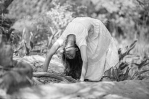A woman in a white dress performing a yoga pose outdoors in a black and white forest setting.