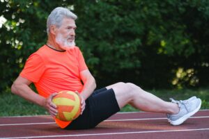 Side view of active senior man with bread in sportswear doing fitness exercise with ball in daytime