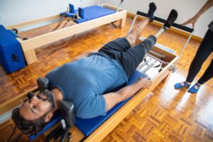 Amputee man in a blue shirt doing physical therapy with a prosthetic leg on Pilates equipment indoors.