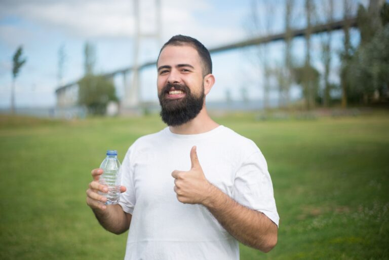 Cheerful man with a beard giving thumbs up while holding a water bottle in a park setting in Portugal.