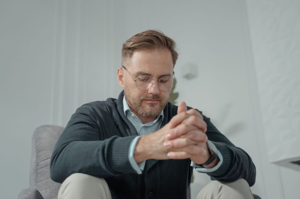 A thoughtful man with glasses and gray sweater sitting indoors, hands clasped.