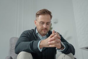 A thoughtful man with glasses and gray sweater sitting indoors, hands clasped.