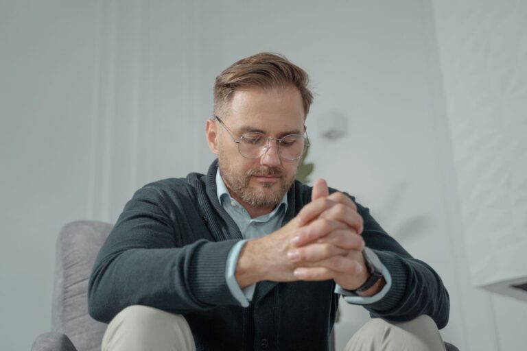 A thoughtful man with glasses and gray sweater sitting indoors, hands clasped.