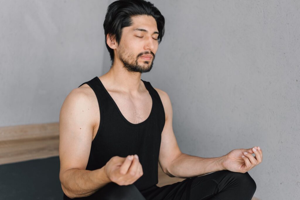 A young man with a beard meditates indoors, eyes closed, in a peaceful lotus pose.