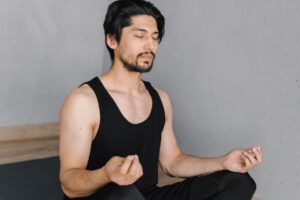 A young man with a beard meditates indoors, eyes closed, in a peaceful lotus pose.
