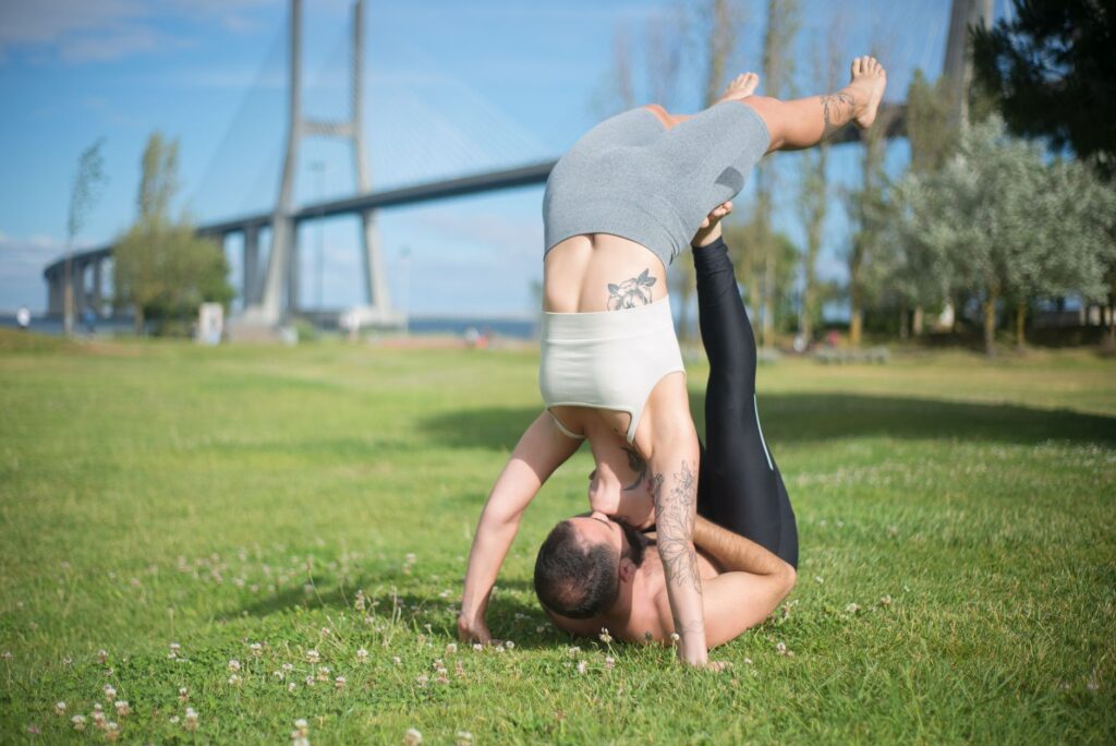 A couple enjoying acrobatic yoga on grass under Lisbon's famous bridge, embracing a healthy lifestyle.