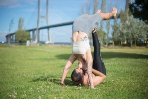 A couple enjoying acrobatic yoga on grass under Lisbon's famous bridge, embracing a healthy lifestyle.
